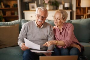 An elderly couple, sitting on a sofa, reviews paperwork and a laptop